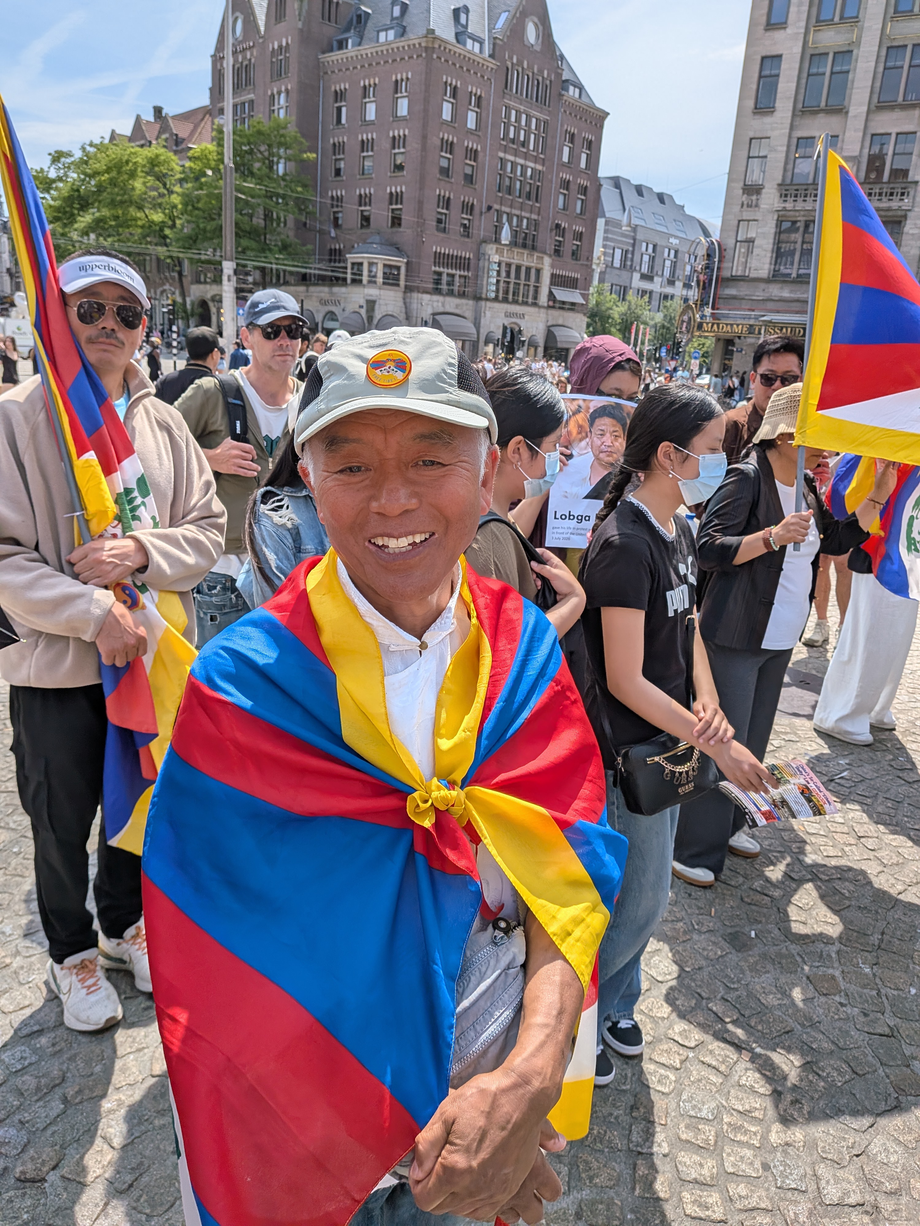 Een man gehuld in de Tibetaanse vlag glimlacht tussen de deelnemers op de Dam