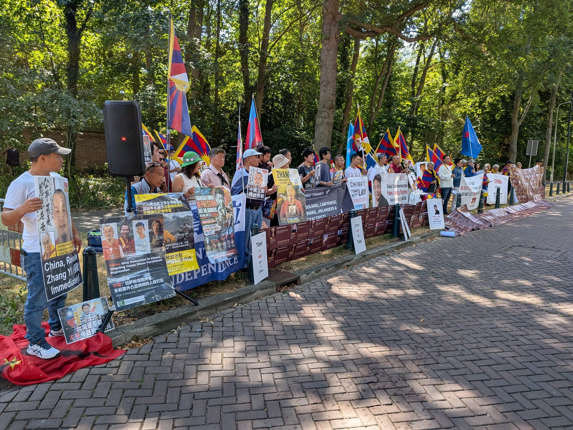 Rij demonstranten langs de Adriaan Goekooplaan met borden, portretten en vlaggen, met een geluidsinstallatie en een Chinese vlag op de grond