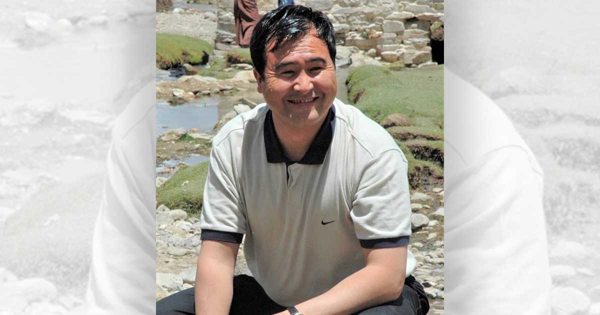 Portret van Dorjee Tashi, glimlachend in een lichte polo, zittend in een Tibetaans rivierlandschap met stenen en groene oevers