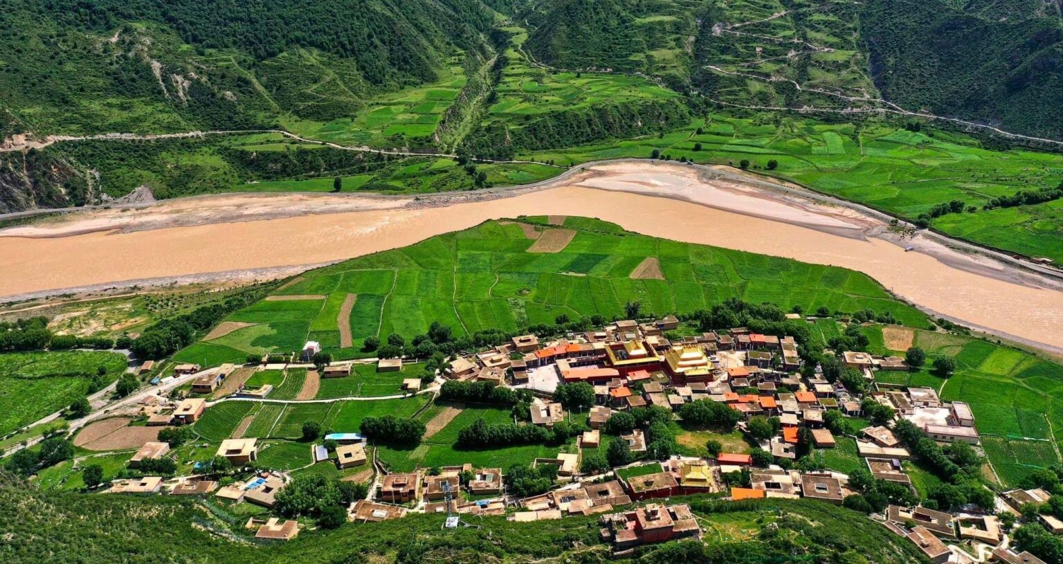 Luchtfoto van het Yena-klooster met gouden daken, omringd door groene akkers en dorpshuizen aan de bruine Drichu-rivier in een bergdal