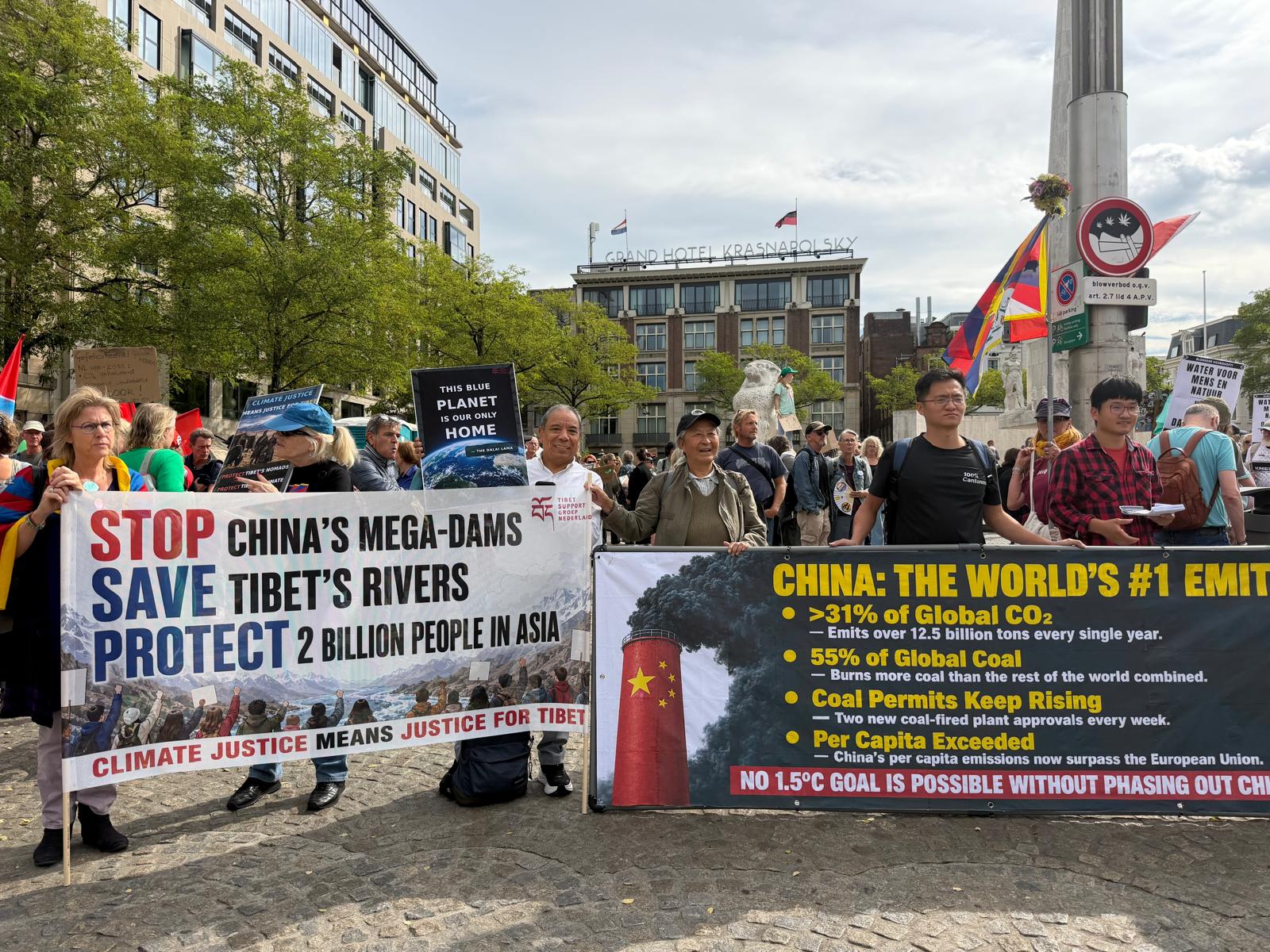 TSG-deelnemers op de Dam met grote spandoeken over China's megadammen en CO2-uitstoot, omringd door Tibetaanse vlaggen bij het Nationaal Monument